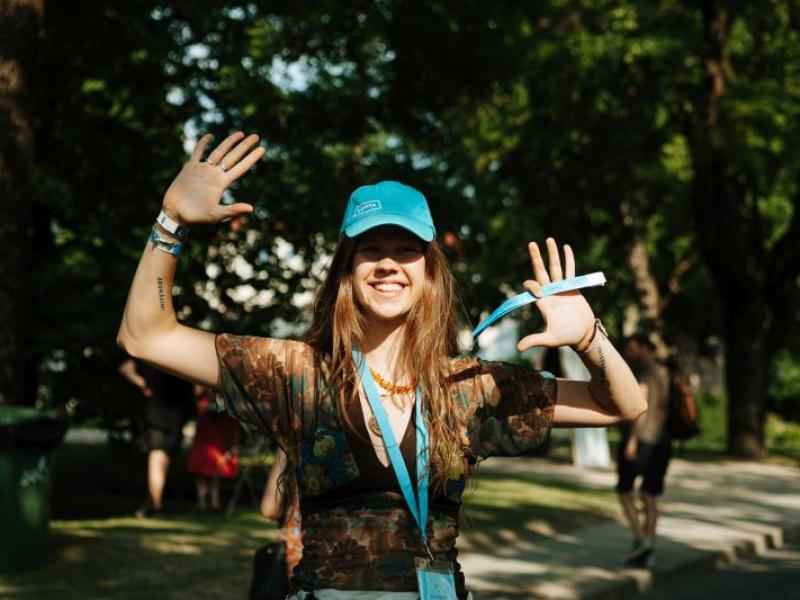 Smaidošs cilvēks zilā cepurē priecīgi paceļ rokas saulainā parkā. Viņam mugurā ir ziedu raksta krekls, kas rada bezrūpīgu noskaņu.