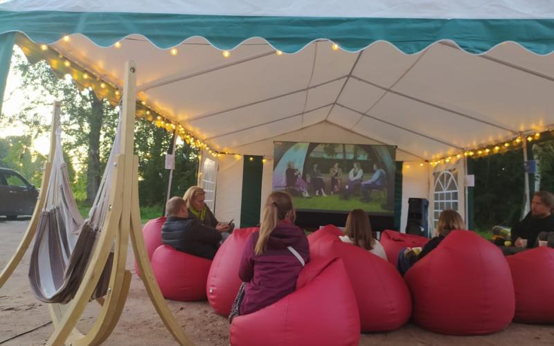 Mājīga brīvdabas filmu seanss zem telts ar virtenēm. Cilvēki atpūšas sarkanos pupu maisos, skatoties pret ekrānu, radot siltu, kopīgu atmosfēru.