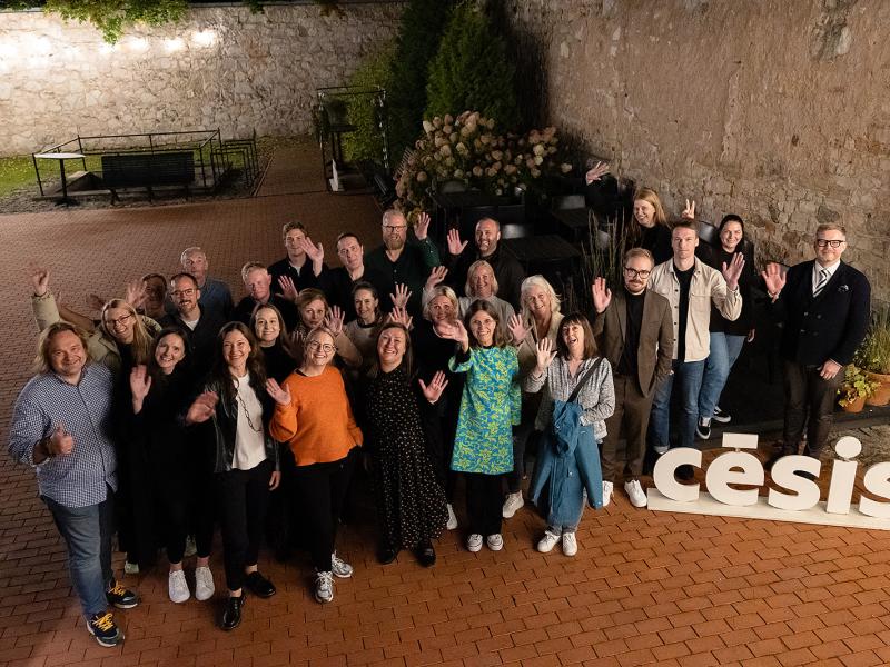 Smaidīgu cilvēku grupa māj kamerai ārā uz ķieģeļu terases, uz zemes lieliem burtiem rakstīts "Cēsis".