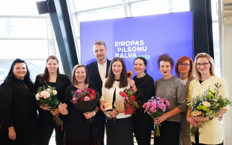 Deviņu cilvēku grupa smaida iekštelpās, rokās turot ziedus. Fonā esoša zīme ar uzrakstu "Eiropas Pilsoņu Balva 2023", kas norāda uz apbalvošanas pasākumu.