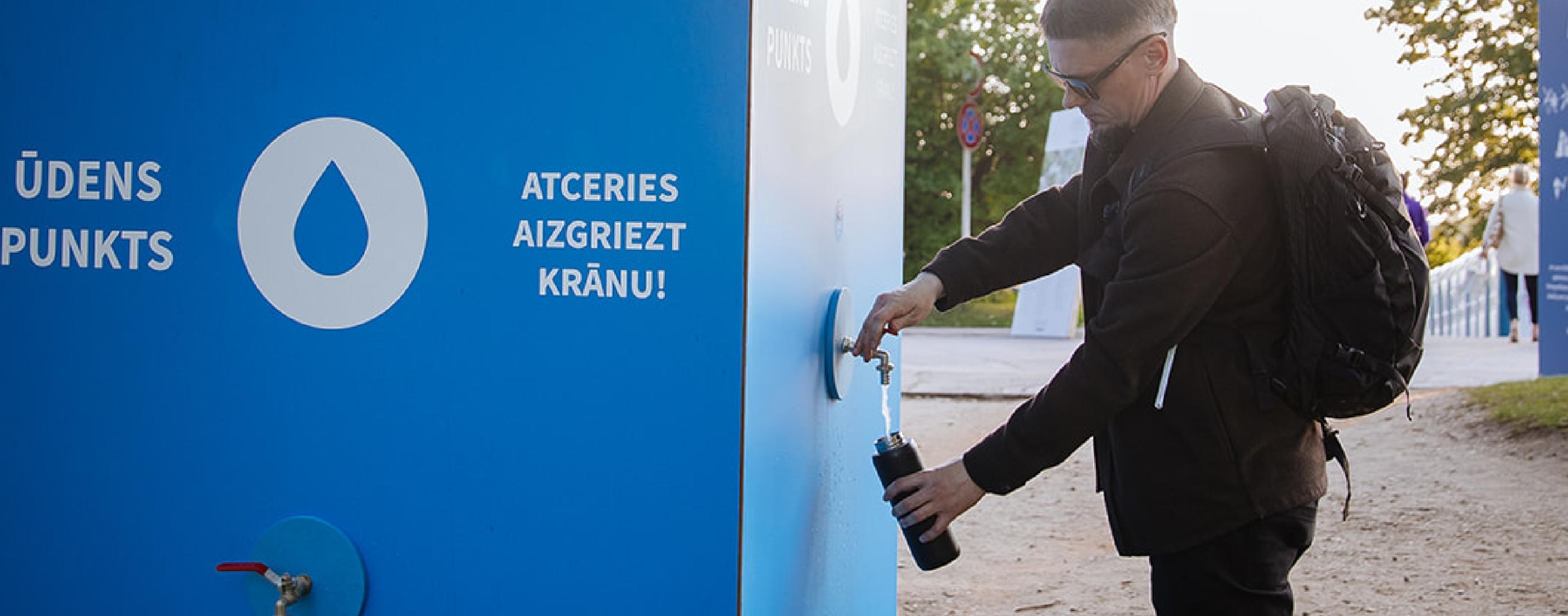 Vīrietis pilda pudeli zilā ūdens uzpildes stacijā ārpus telpām. Zīme uz uzpildes stacijas norāda lietotājiem aizgriezt krānu. Aina ir labi apgaismota un mierīga.