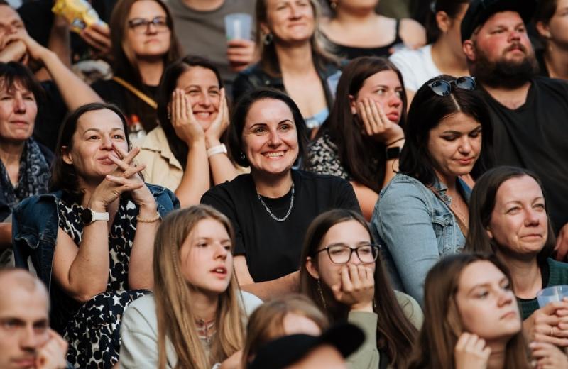 Liels, daudzveidīgs pūlis sēž cieši saspiedies, uzmanīgi vērojot ar ieinteresētām un smaidošām sejām. Atmosfēra ir dzīvīga un aizraujoša.