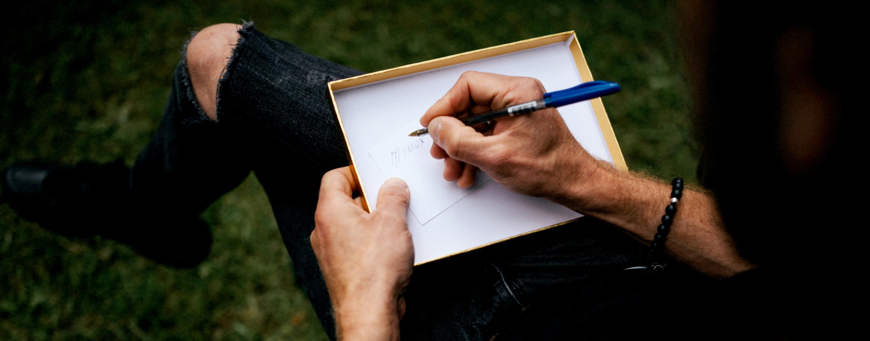 Cilvēks melnā apģērbā raksta uz papīra lapas, kas novietota uz ceļiem, izmantojot zilu pildspalvu. Viņš sēž ārā uz zāles.