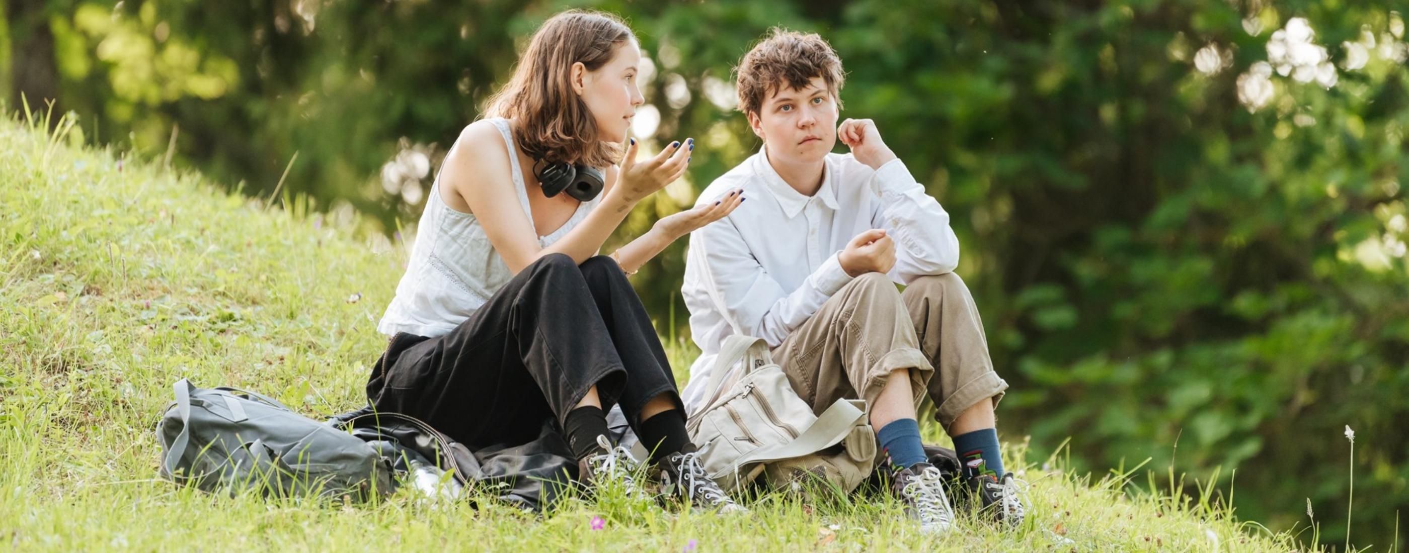 Divi jaunieši sēž zālē, iesaistījušies sarunā. Viens izteiksmīgi žestikulē, kamēr otrs uzmanīgi klausās. Fonā koki