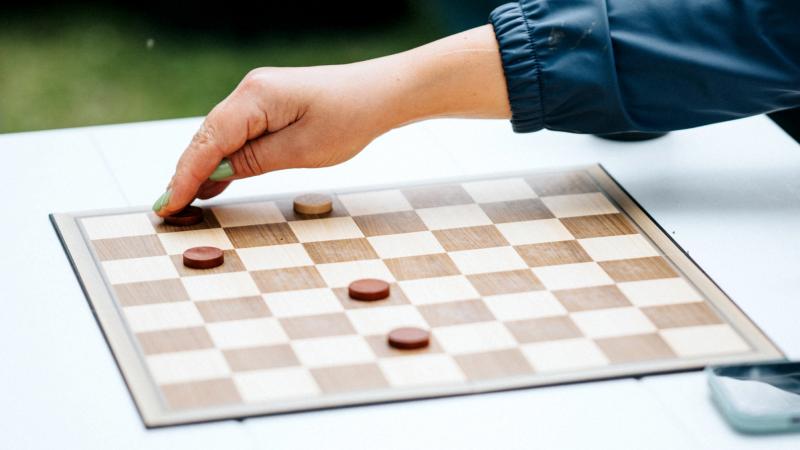 Roka zilā piedurknē pārvieto figūru uz dambretes galdiņa, redzamas piecas figūriņas. Aina atrodas ārā, radot relaksētu, stratēģisku atmosfēru.