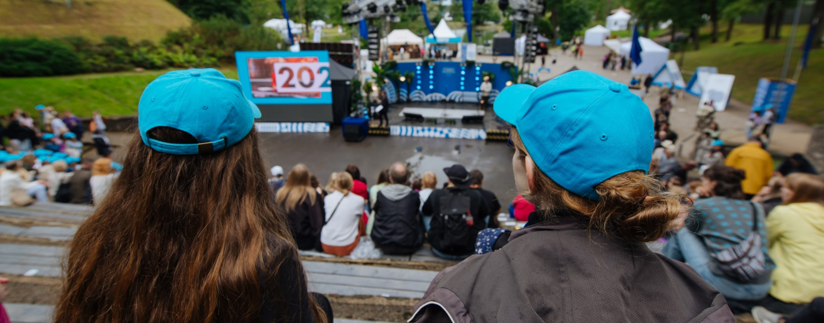 Divi cilvēki zilās cepurēs vēro pasākumu brīvā dabā no pārpildītas auditorijas aizmugures.