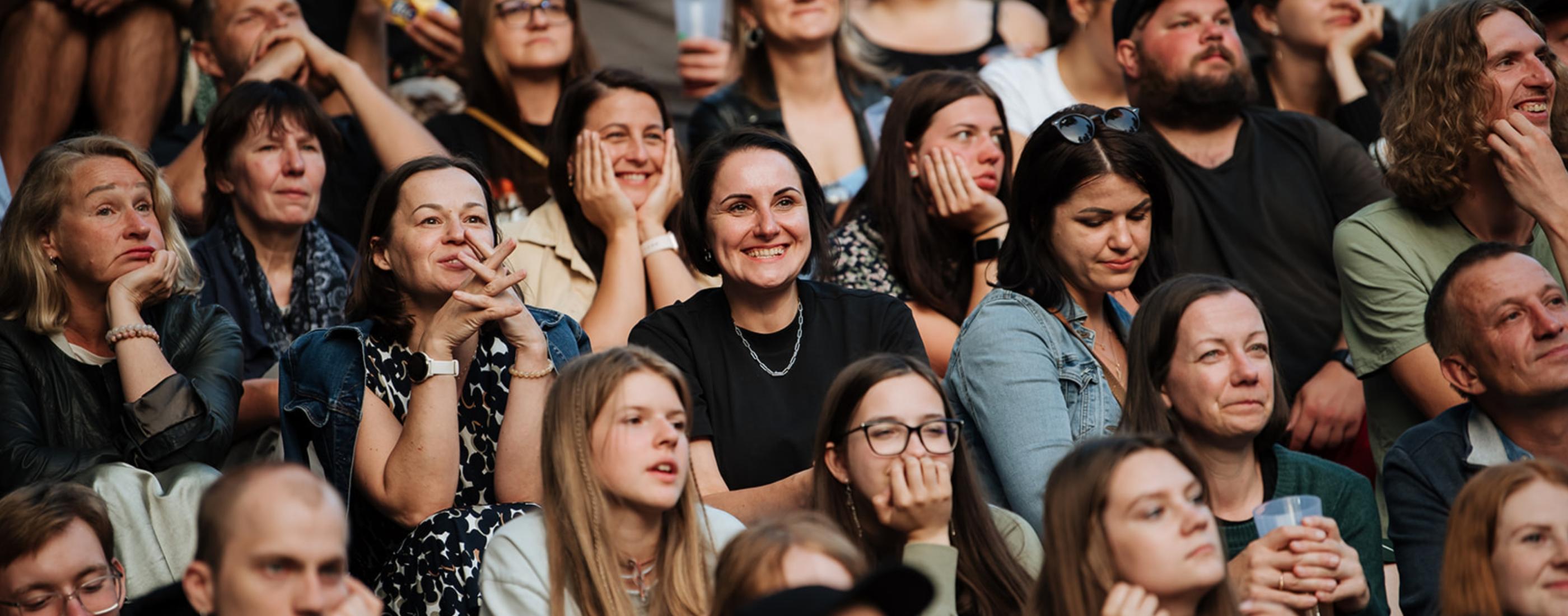 Liels, daudzveidīgs pūlis sēž cieši saspiedies, uzmanīgi vērojot ar ieinteresētām un smaidošām sejām. Atmosfēra ir dzīvīga un aizraujoša.
