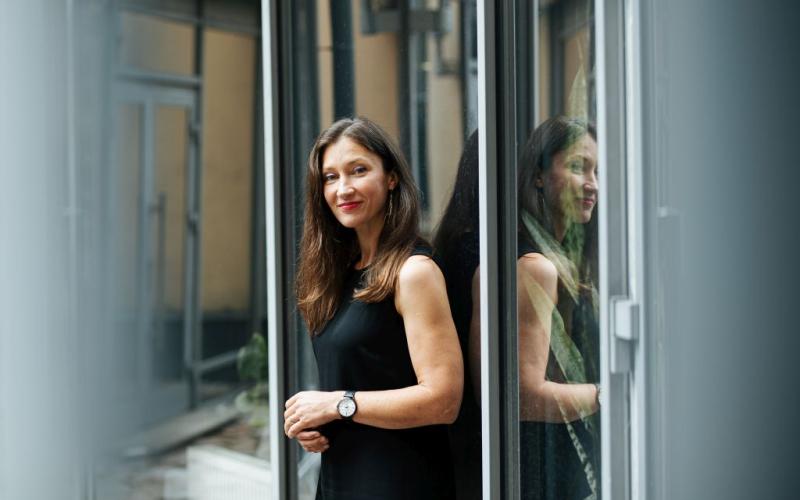 Ieva Morica melnā kleitā stāv pie stikla sienas, un viņas atspulgs ir redzams. Viņa maigi smaida, paužot mierīgu un pārliecinātu smaidu.