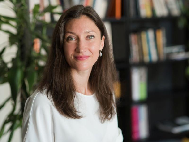 Ieva Morica baltā kreklā maigi smaida. Viņa sēž istabā, kuras fonā ir augs un izplūdis grāmatu plaukts.