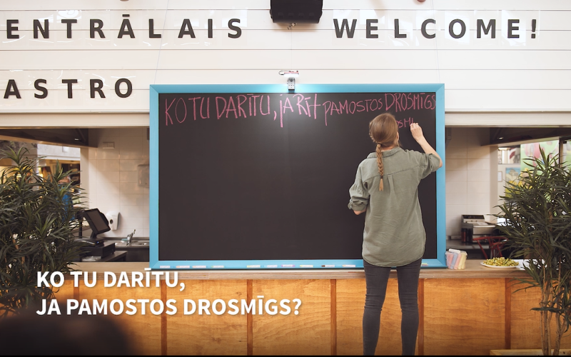 Kafejnīcā cilvēks raksta uz tāfeles.  Priekšplānā teksts: "Kotu darītu, ja pamostos drosmīgs?".Ainu ierāmē augi, radot viesmīlīgu atmosfēru.