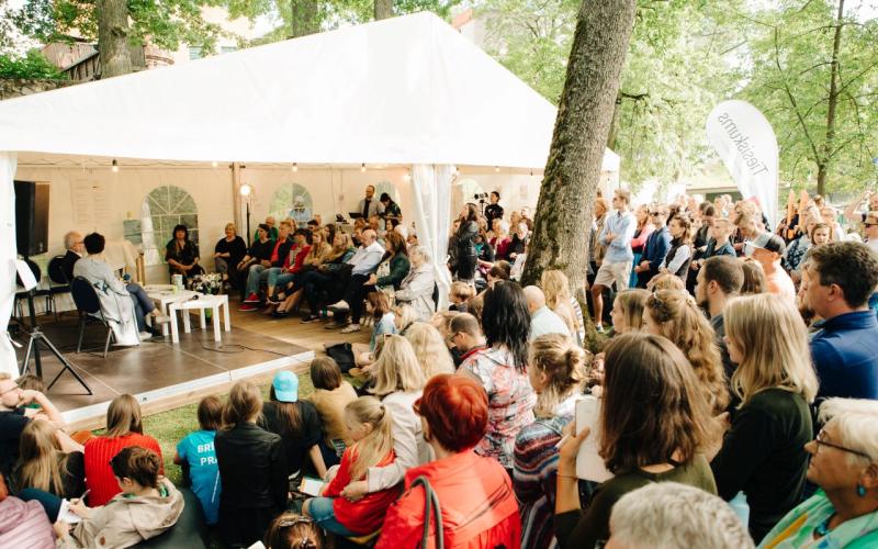 Liela cilvēku grupa uzmanīgi vēro paneļdiskusiju zem baltas telts zaļā parkā. Atmosfēra ir dzīvīga un iesaistīta.