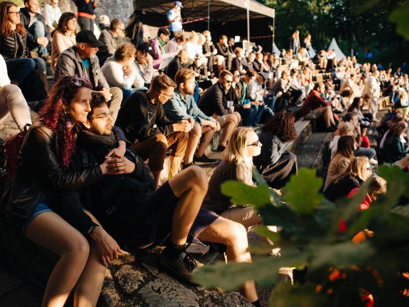 Dažādu cilvēku pūlis sēž uz akmens kāpnēm brīvā dabā, baudot saulainu pasākumu. Atmosfēra ir relaksēta un dzīvespriecīga, un tērpi ir ikdienišķi.