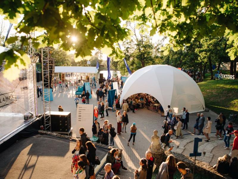 Dinamisks brīvdabas festivāls, kurā pūļi pulcējas ap lielu, baltu telti zem lapu kokiem. Spoža saules gaisma met garas ēnas, radot dzīvīgu un dzīvespriecīgu atmosfēru.