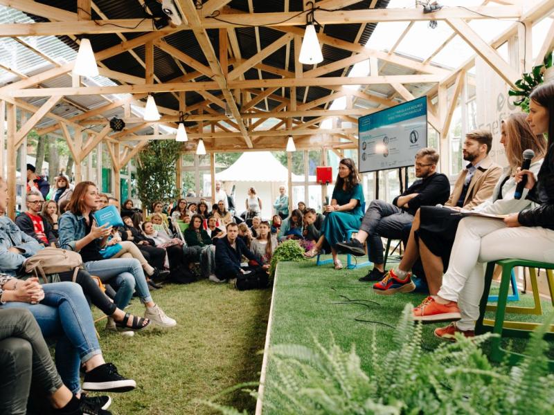 Piecu cilvēku panelis sēž uz skatuves ar mikrofoniem, iesaistot sēdošu auditoriju siltumnīcai līdzīgā struktūrā. Tonis ir dzīvīgs un interaktīvs.