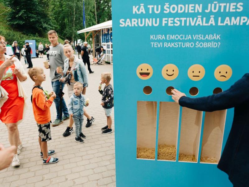 Cilvēku grupa stāv pie tirkīzzilas atsauksmju tāfeles ar dažādiem emocijzīmju simboliem emociju izvēlei.