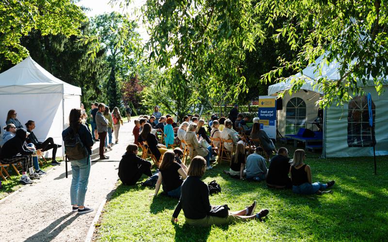 Pulcēšanās brīvā dabā saulainā parkā, kur cilvēki sēž uz krēsliem un zālē, klausoties runātājā pie baltas telts. Koki rada mierīgu fonu.