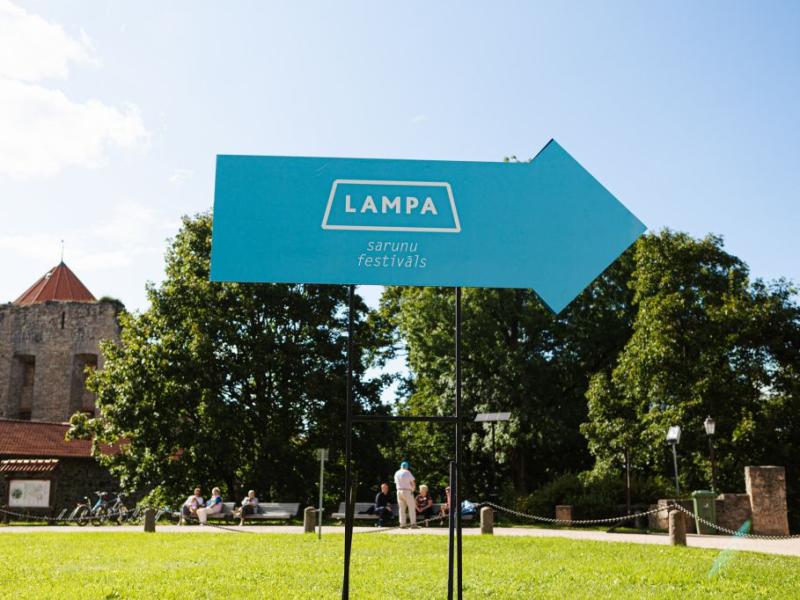 Zila bultiņas formas zīme ar zīmi, kas norāda pa labi, atrodas uz koku un senas akmens ēkas fona zem skaidrām debesīm.