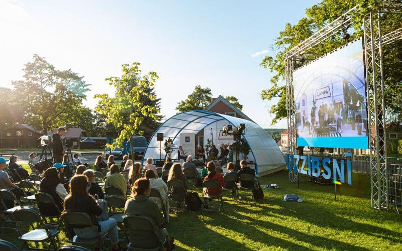 Āra pasākums saulrietā parkā ar sēdošu auditoriju, kas vērsta pret skatuvi kupola teltī. Pasākums tiek rādīts uz liela ekrāna. Apkārtni ieskauj koki.