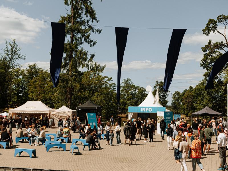Āra pasākums, kurā cilvēki pulcējas ap teltīm ar uzrakstu "INFO". Zili soliņi un karogi rada dzīvīgu, svētku atmosfēru zem skaidrām debesīm.