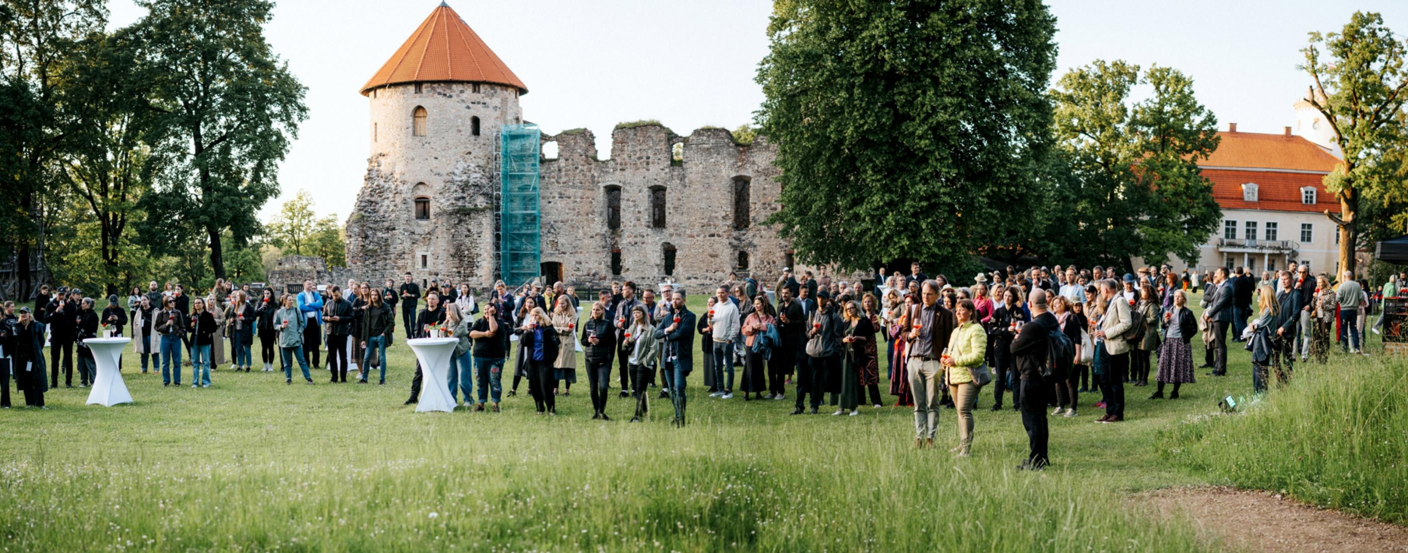 Liels pūlis pulcējas zālienā netālu no vēsturiskās Cēsu pils pilsdrupām. Teritoriju ieskauj koki, radot dzīvīgu, svētku atmosfēru.