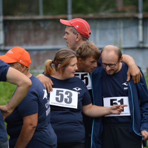 Cilvēku grupa sporta sacensību laikā, visi ģērbušies vienādos sporta kreklos ar numuriem, cits citu atbalsta un apskauj, izrādot komandas garu un draudzību.